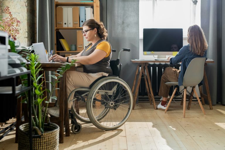 Young disabled businesswoman in casualwear is sitting in a wheelchair in front of a laptop typing while her colleague works at her computer near the window across the room